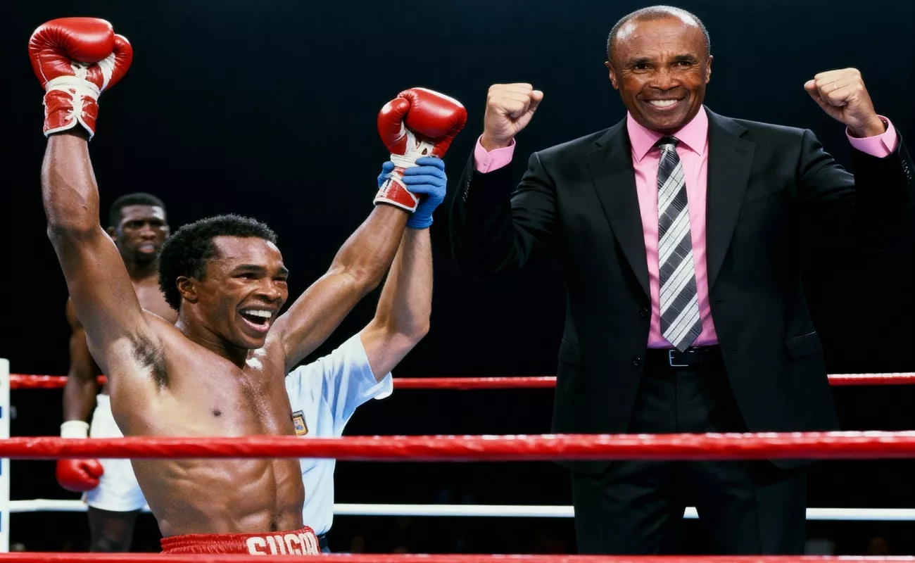 Bokser triumfalnie unosi ręce w rękawicach, stojąc na ringu bokserskim. Obok niego uśmiechnięty mężczyzna w garniturze również podnosi pięści w geście zwycięstwa.