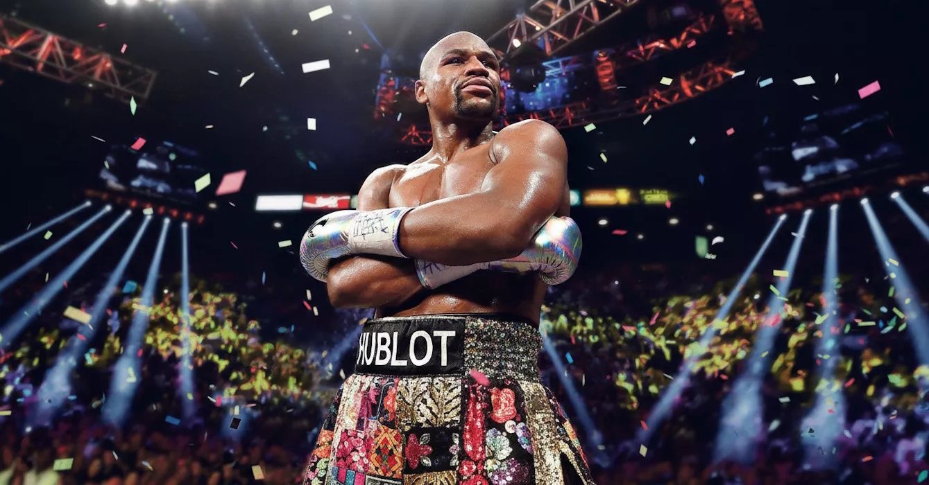 Bokser stoi z założonymi rękami, w srebrnych rękawicach. Na jego szortach widnieje napis "HUBLOT". W tle widać trybuny pełne ludzi i spadające konfetti.