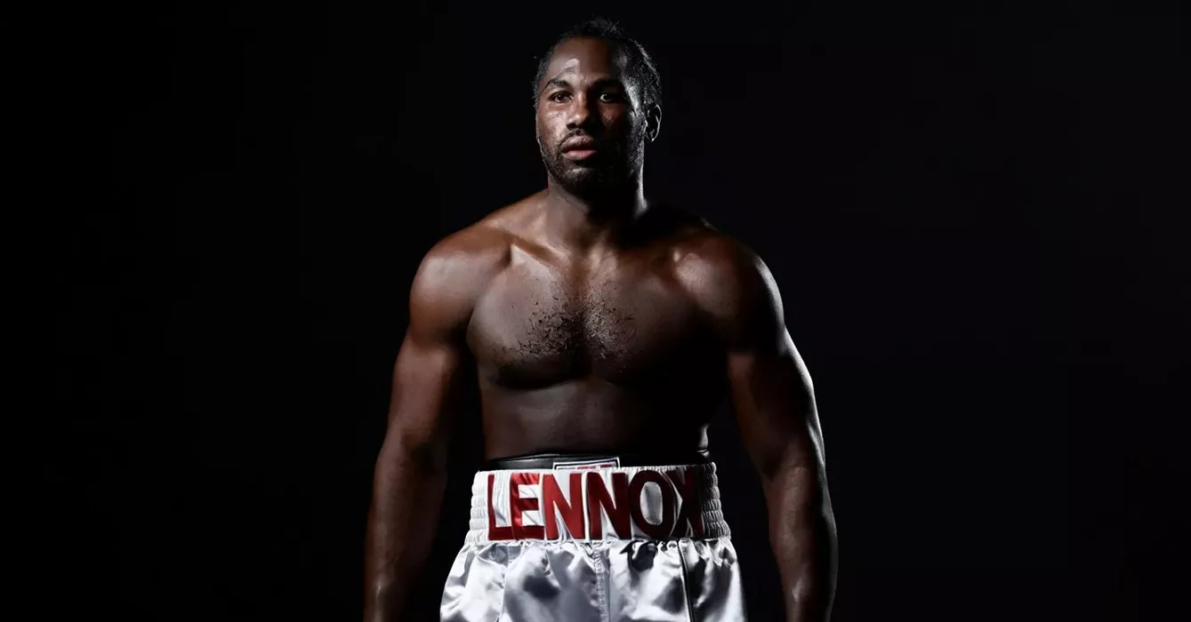 Bokser stoi spokojnie, ubrany w białe szorty z przodu z napisem "LENNOX", na ciemnym tle. Jego umięśnione ciało jest oświetlone, podkreślając atletyczną sylwetkę.