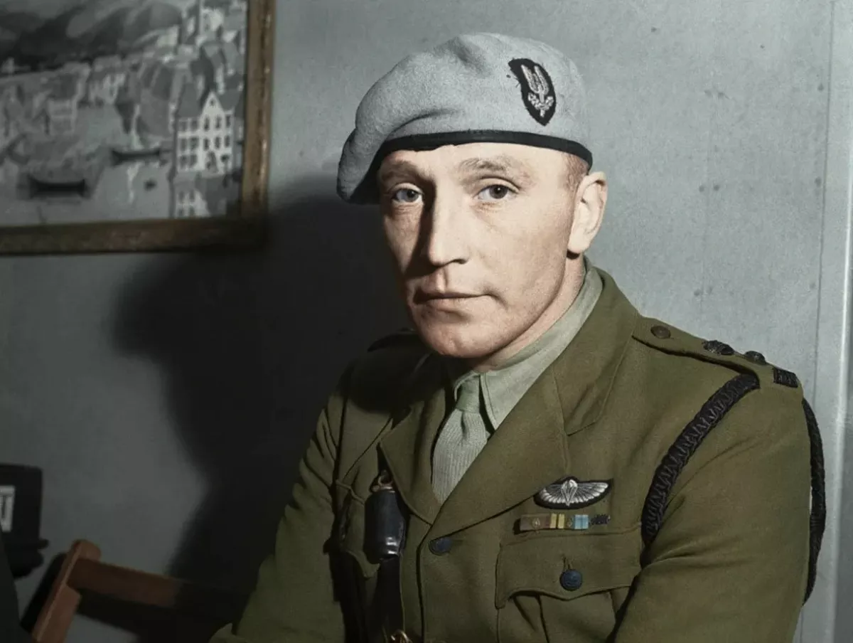 Mężczyzna w mundurze siedzi spokojnie, nosi beret z ozdobą. W tle obraz przedstawiający budynki nad wodą. Ściany są szare, a obok stoi krzesło.