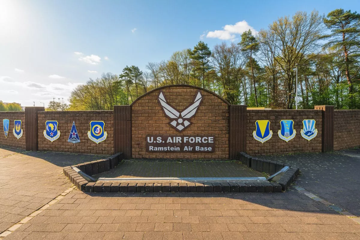 Znak przedstawia logo U.S. Air Force oraz napis "U.S. AIR FORCE Ramstein Air Base" na tle ceglanej ściany z emblematami wojskowymi. W tle widać drzewa i niebo.