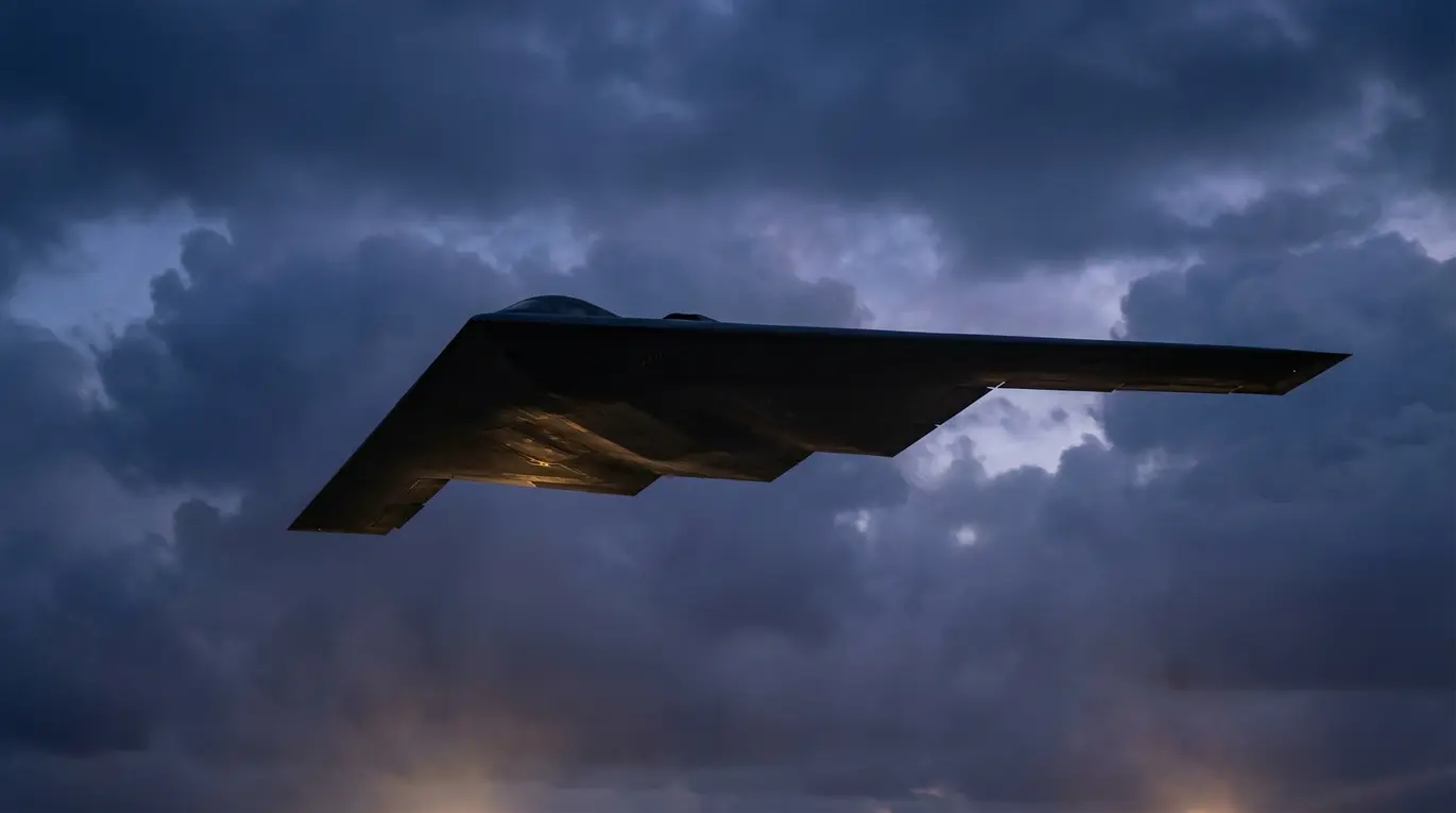 Samolot o kształcie skrzydła przelatuje nad chmurami. Oświetlony jest od dołu, a otaczające go niebo jest ciemne i pochmurne, tworząc dramatyczną atmosferę.