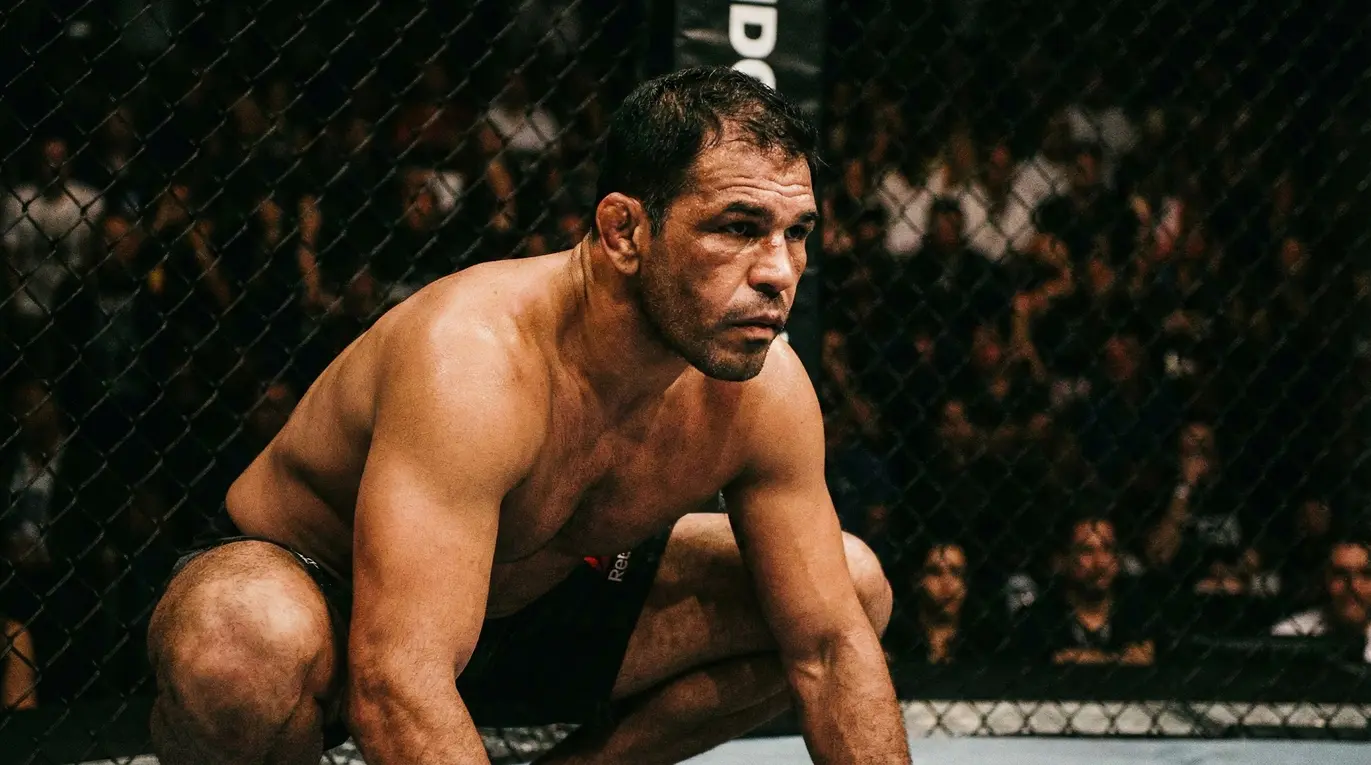 Zawodnik w szortach kuca w klatce, przygotowując się do walki. W tle widać tłum ludzi obserwujących wydarzenie.