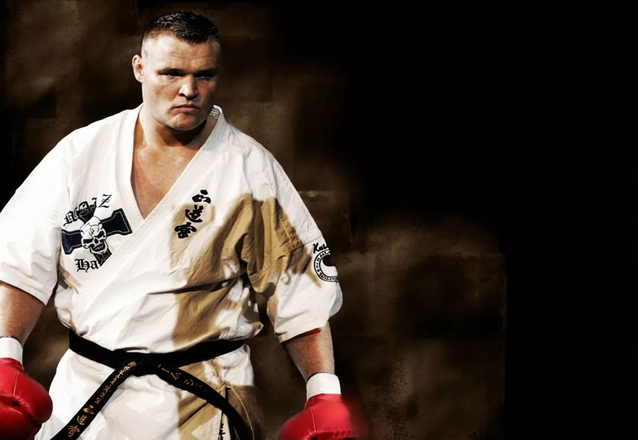 Mężczyzna w gi i czerwonych rękawicach bokserskich stoi w bojowej postawie. Gi zdobione jest japońskimi znakami i grafiką czaszki. Tło jest ciemne i jednolite.