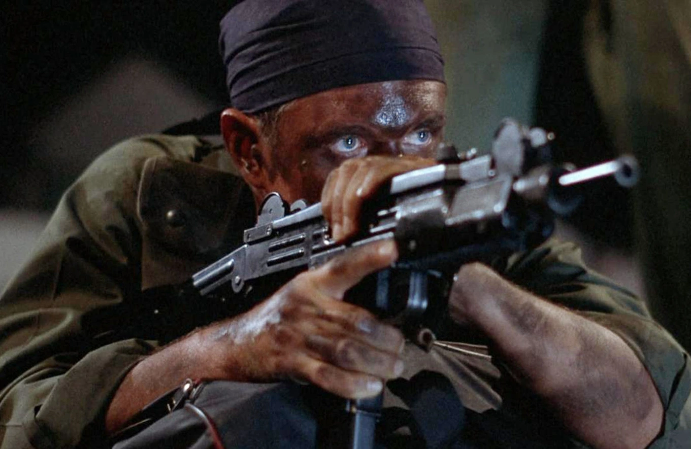 Mężczyzna celuje z karabinu, skupiony z zakamuflowaną twarzą i błękitnymi oczami. Otoczenie jest zaciemnione, sugerując akcję nocną lub w ukryciu.