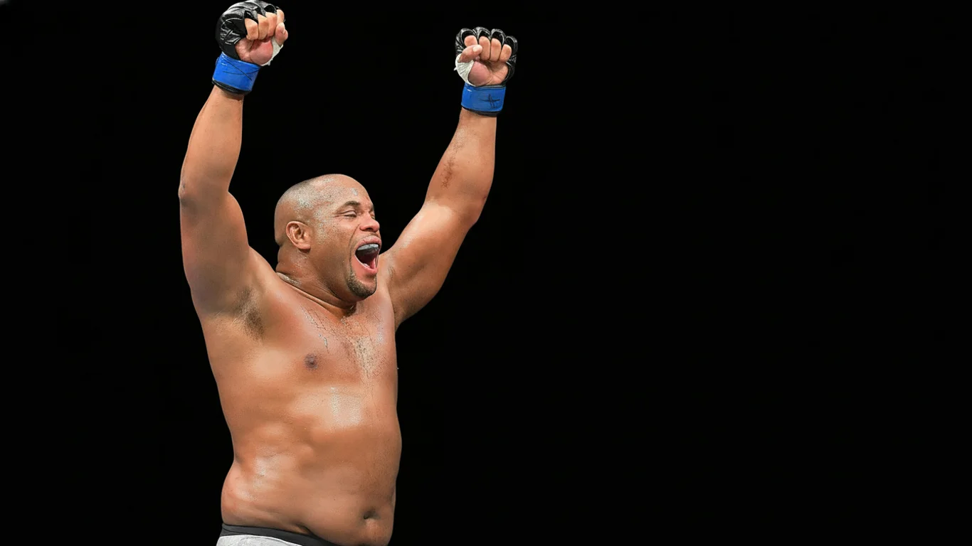 Mężczyzna z podniesionymi ramionami w geście triumfu, uśmiecha się szeroko na tle jednolitego, ciemnego tła. Na jego rękach widoczne są rękawice sportowe.