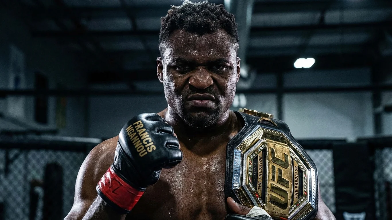 Mężczyzna z napiętą twarzą unosi zaciśniętą pięść z napisem "Francis Ngannou" na rękawicy, trzymając złoty pas UFC, w tle siłownia.