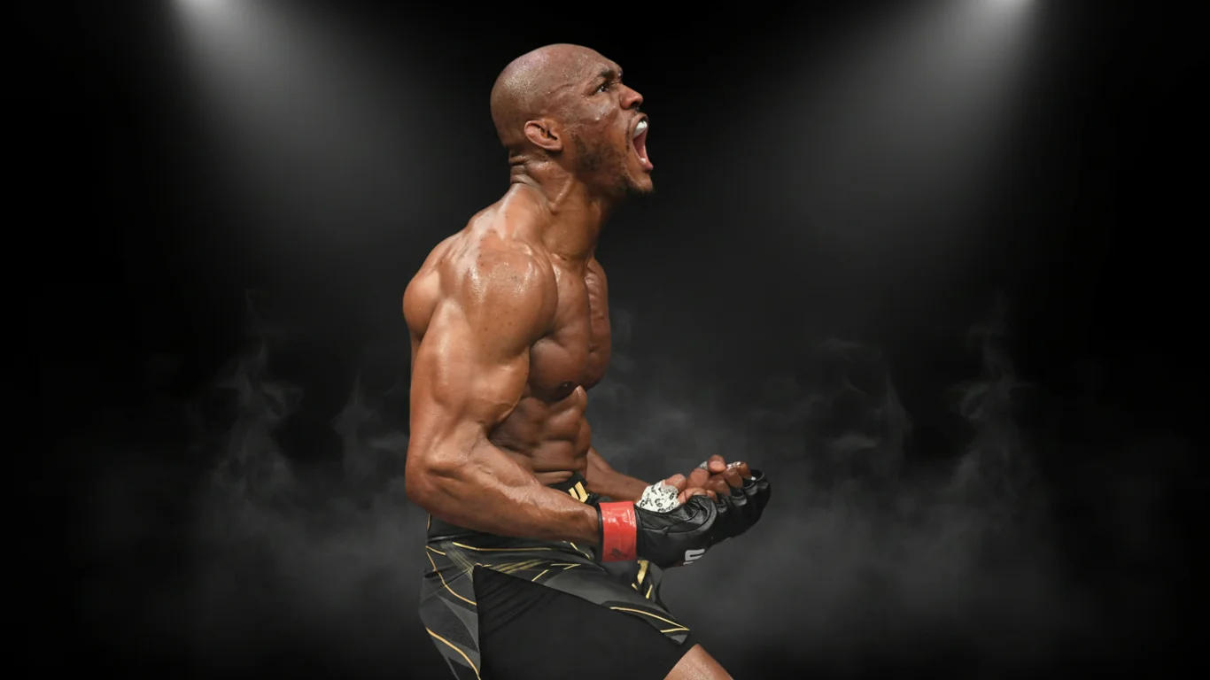 Mężczyzna w sportowym stroju krzyczy, pokazując napięte mięśnie. Stoi na ciemnym tle z reflektorami oświetlającymi go z góry.