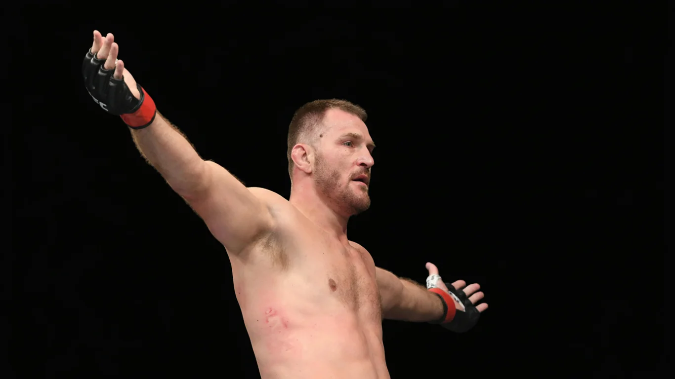 Mężczyzna z rękawicami UFC rozkłada ręce, stojąc na czarnym tle, ukazując umięśnioną sylwetkę i wyraz determinacji na twarzy.