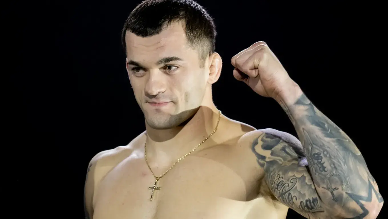 Mężczyzna z krótkimi włosami, w naszyjniku z krzyżykiem, podnosi zaciśniętą pięść. Widoczne są tatuaże na ręce. Tło jest czarne, co podkreśla jego sylwetkę.