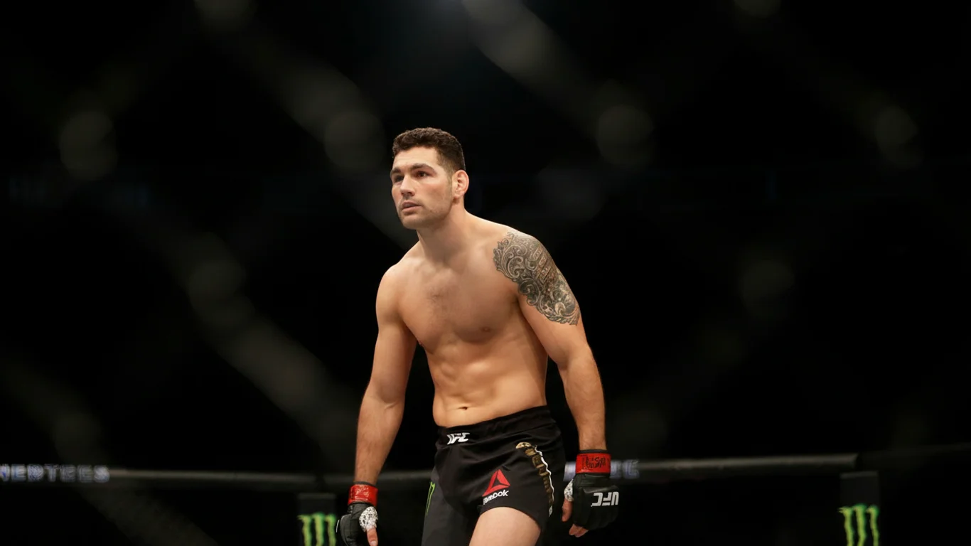 Zawodnik MMA w czarnych spodenkach stoi w pozycji gotowości w oktagonie. Na lewym ramieniu widoczny jest tatuaż. Tło jest ciemne, rozmyte.