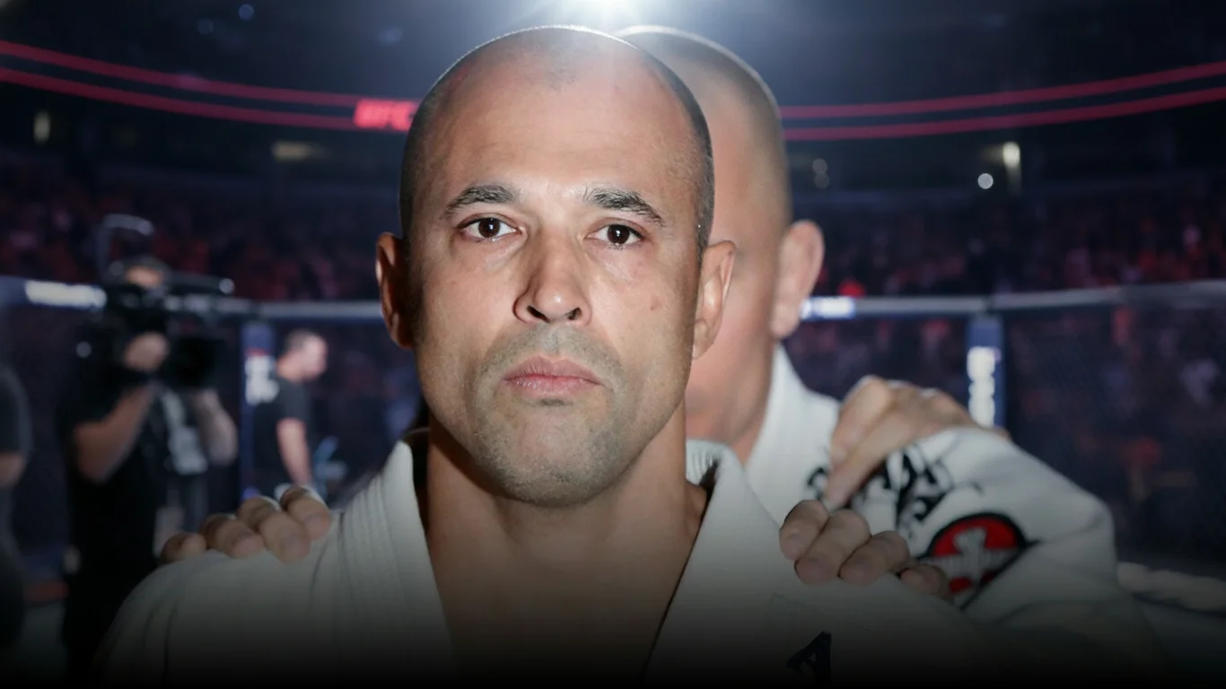 Mężczyzna w stroju sportowym stoi z poważnym wyrazem twarzy, trzymając się za ramiona. W tle widać rozmytą scenę areny sportowej.