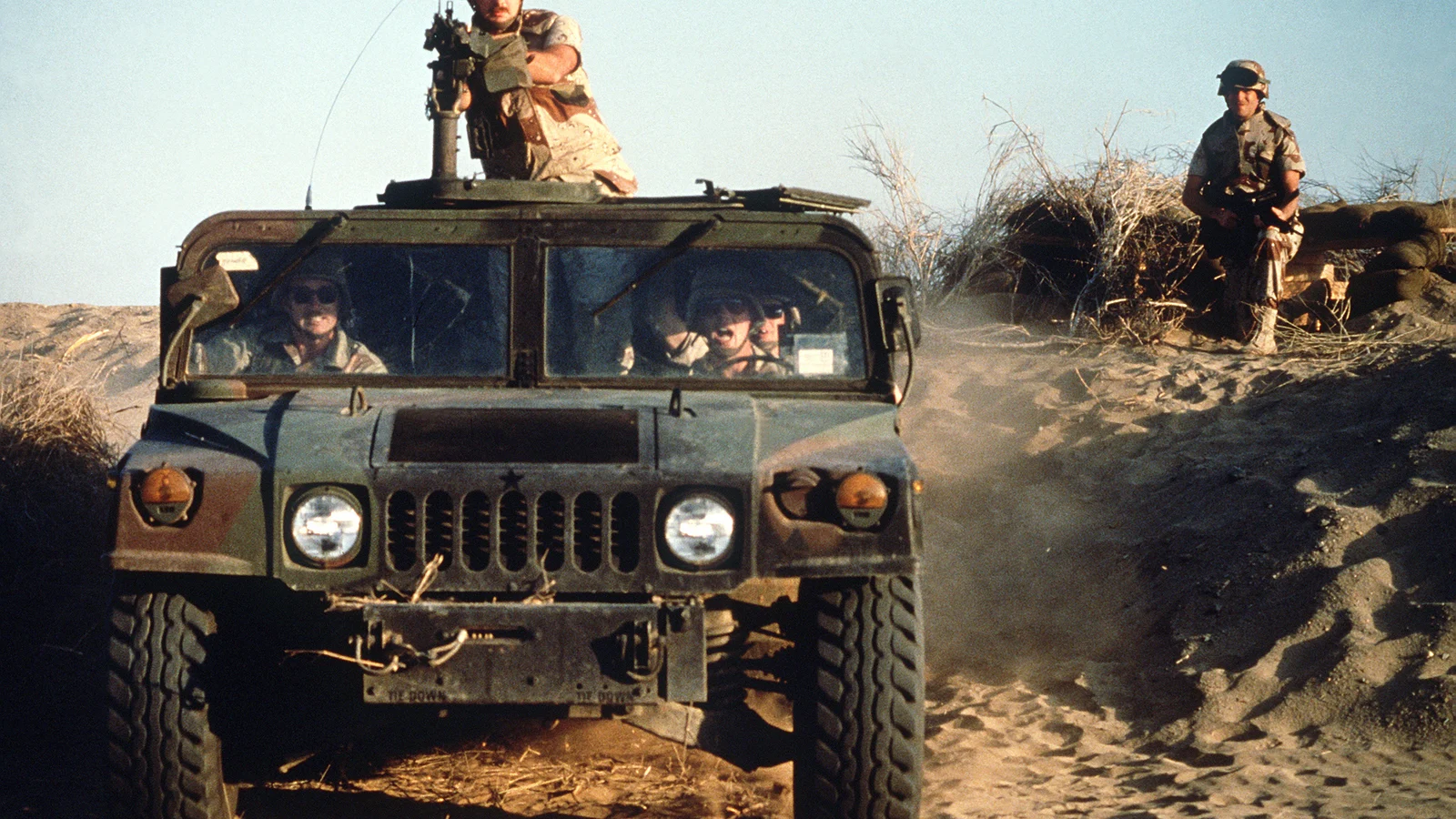 Terenowy M1038 HMMWV w kamuflażu leśnym jadący po piasku, strzelec z karabinem maszynowym na dachu, w tle żołnierz na stanowisku z worków z piaskiem