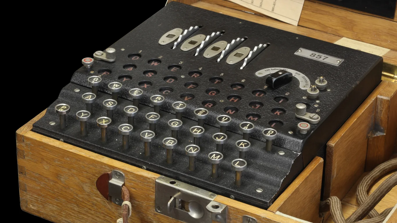 Niemiecka wojskowa maszyna szyfrująca Enigma w drewnianej skrzynce, z klawiaturą, panelem lampek i mechanizmem wirników, na czarnym tle.