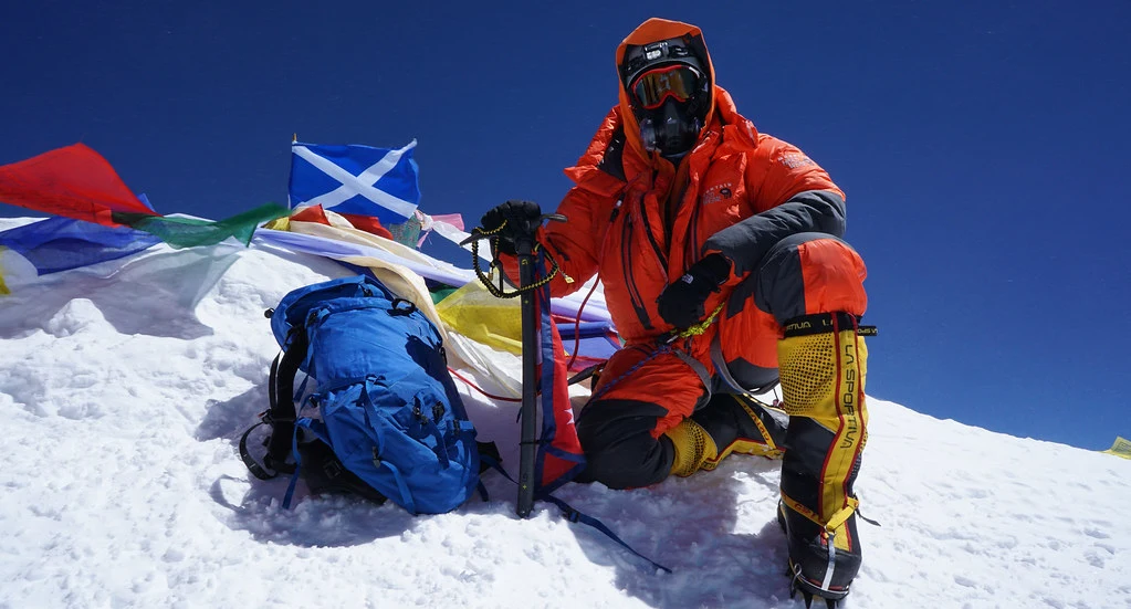 Alpinista w kombinezonie puchowym i masce tlenowej na ośnieżonym szczycie ośmiotysięcznika