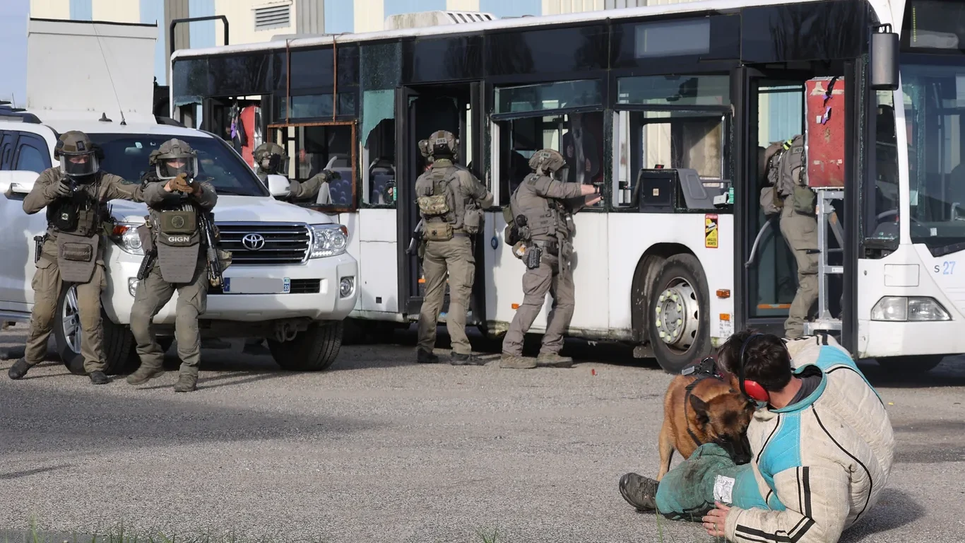Operatorzy GIGN w hełmach i maskach szturmują autobus podczas pokazu odbijania zakładników, w tle pies służbowy obezwładniający pozoranta