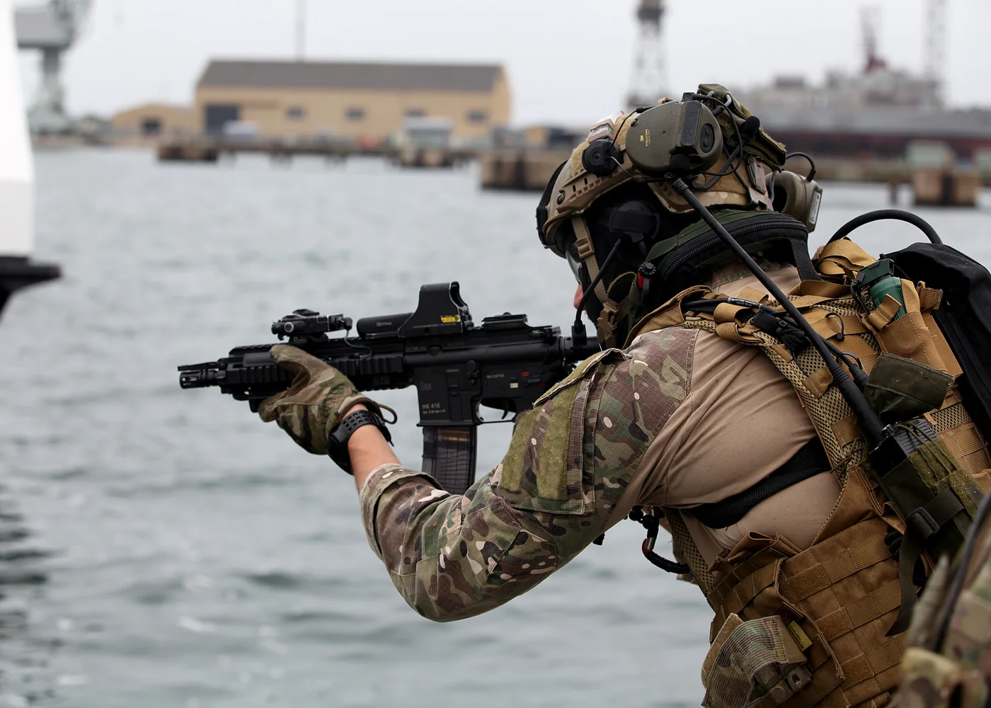 Operator GROM w kamuflażu MultiCam i hełmie z aktywnymi ochronnikami słuchu celuje z karabinka HK416 nad wodą w porcie