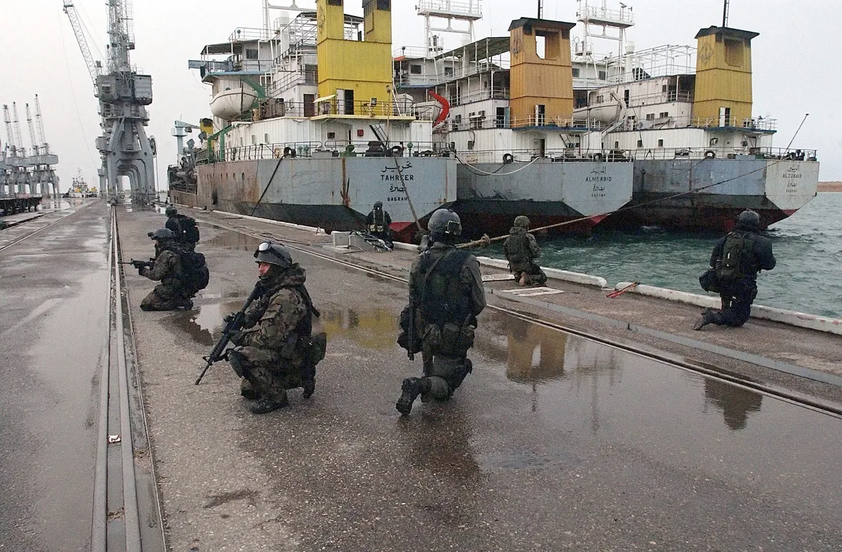 Żołnierze GROM klęczący z bronią na nabrzeżu portowym, w tle irackie statki i dźwigi portowe