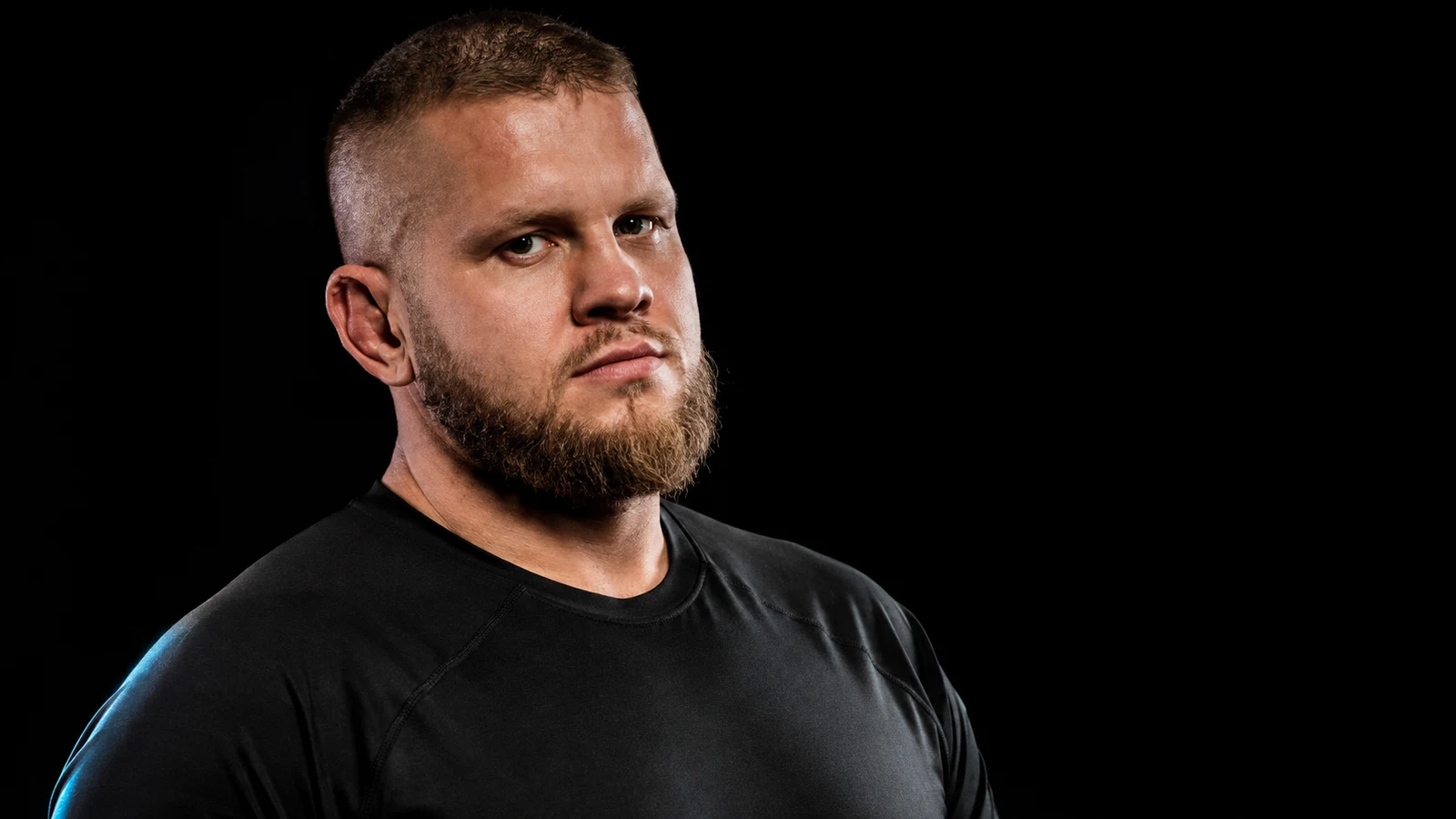 Portret zawodnika MMA wagi ciężkiej z krótko ostrzyżonymi włosami i blond brodą, w czarnej koszulce, na czarnym tle